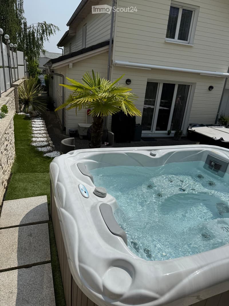Private garden area with a modern hot tub, surrounded by a stone wall, greenery, and a house with large windows and a sliding door.