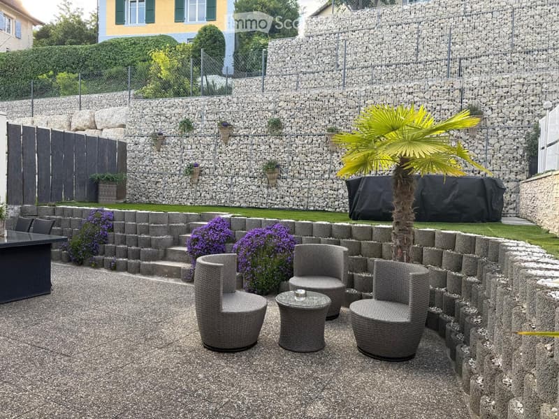 Garden area with a stone wall, seating area with wicker chairs and a table, surrounded by flowers and a palm tree.