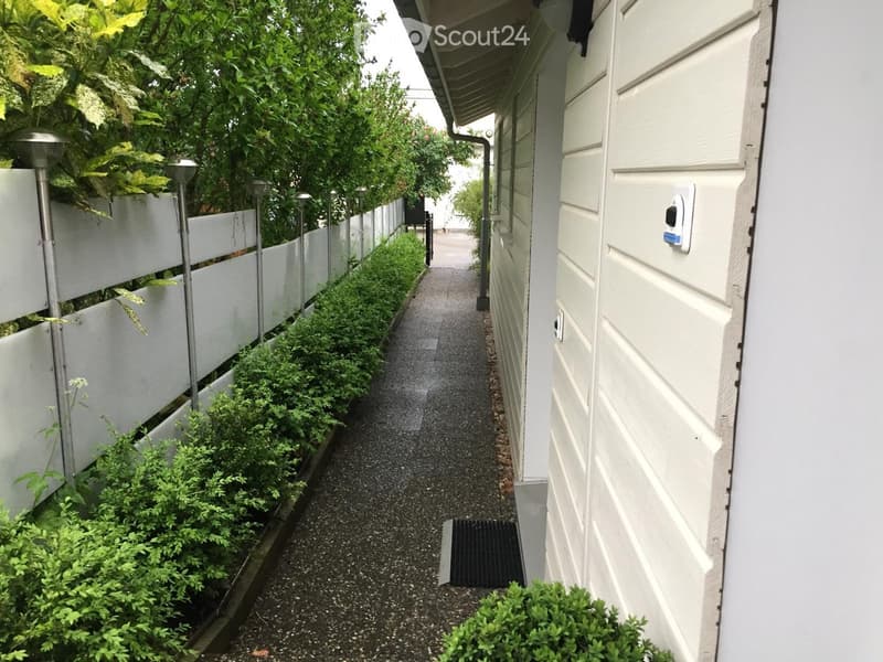 A narrow, gravel-covered pathway bordered by white fences and lush greenery, leading to a building entrance.