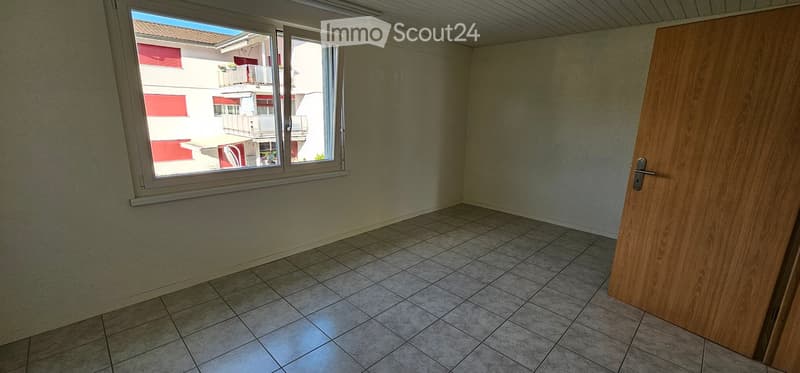 Empty room with tiled floor, white walls, and a large window offering a view of a neighboring building with balconies.