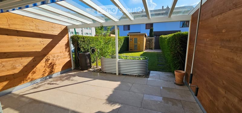 A covered patio area with tiled flooring, wooden walls, and a metal planter in the center. The background shows a well-maintained garden with hedges, a small wooden shed, and a blue house.