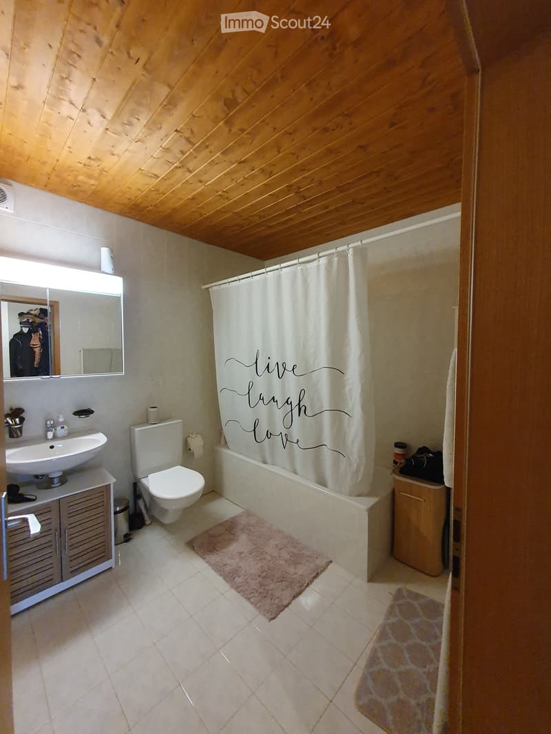 Bathroom with a wooden slanted ceiling, white tiled walls, a sink with a cabinet, a toilet, a bathtub with a shower curtain, and a pink rug on the floor.