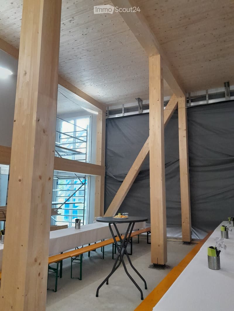 Modern, open-concept space with exposed wooden beams and pillars, featuring a long bench seating area, a small round table, and a bar area with wine glasses and utensils.