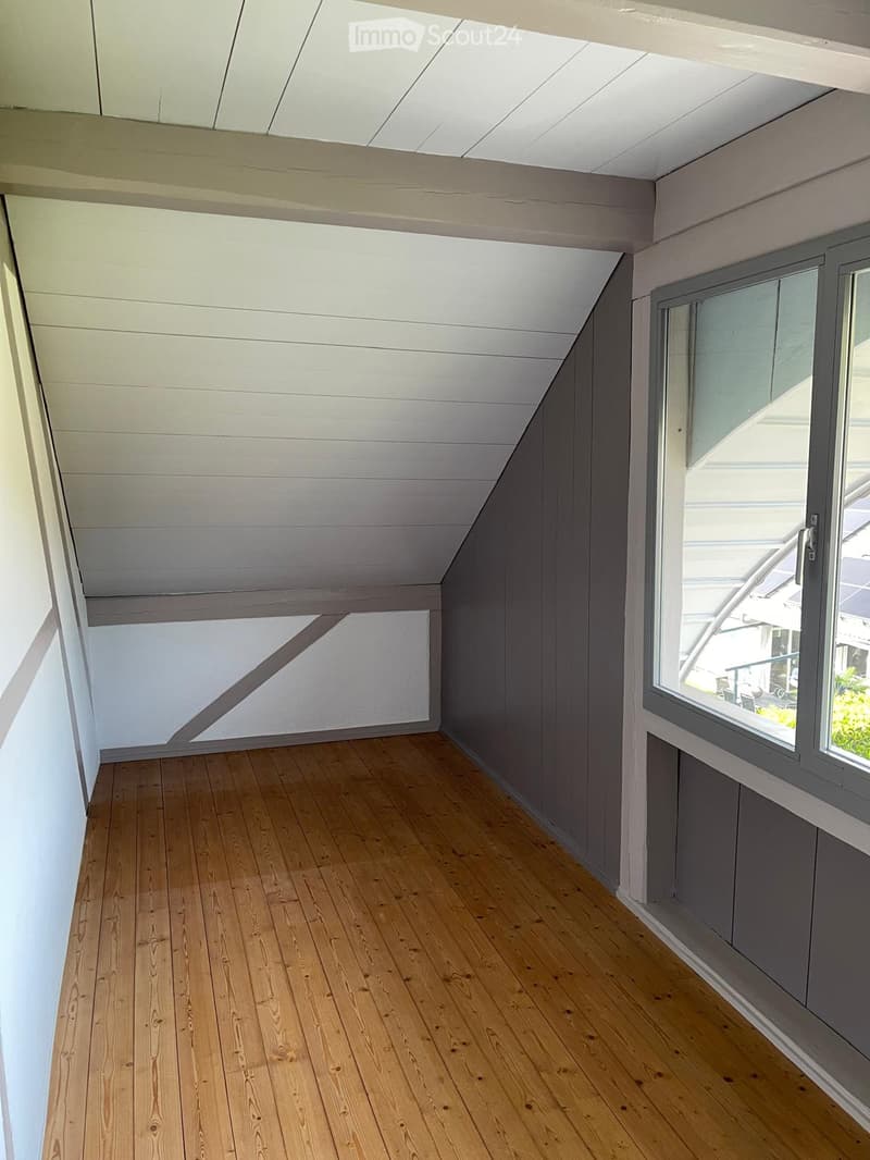 Empty room with wooden flooring, white walls, and large windows allowing natural light. The ceiling has a slanted design with exposed beams.