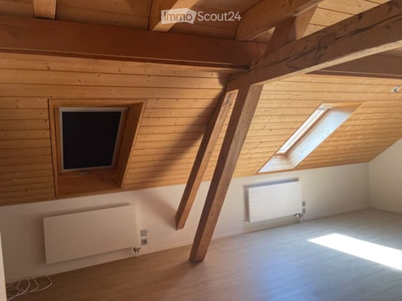 Bright, empty attic room with wooden walls, exposed wooden beams, and skylight windows. Features built-in radiators and a smooth wooden floor.
