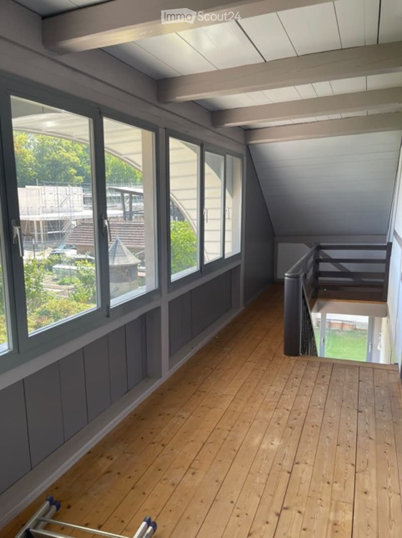 Spacious wooden balcony with large windows, grey walls, and a view of the surrounding area.