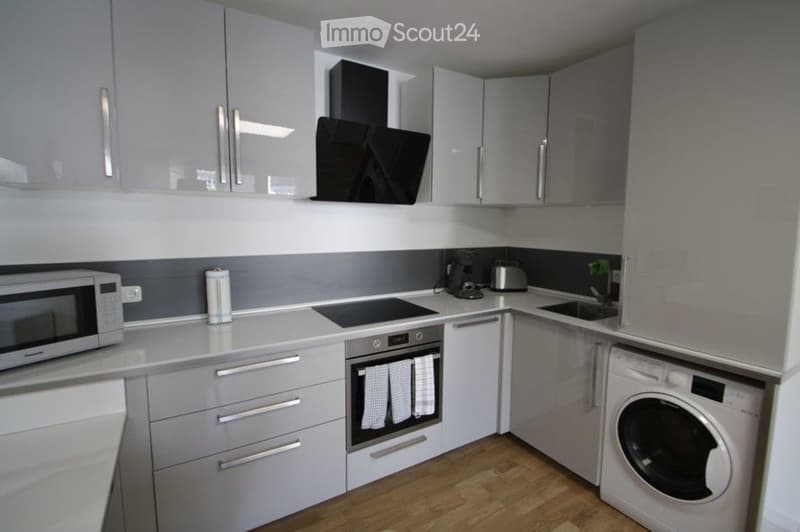 Modern kitchen with grey cabinets, white countertops, and stainless steel appliances including a microwave, coffee maker, toaster, oven, and washing machine. Features a range hood and a sink.
