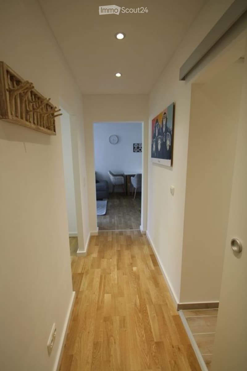 A narrow hallway with wooden flooring, recessed ceiling lights, and a small decorative shelf on the left wall. At the end of the hallway, a doorway leads to a room with a couch, chairs, and a coffee table.