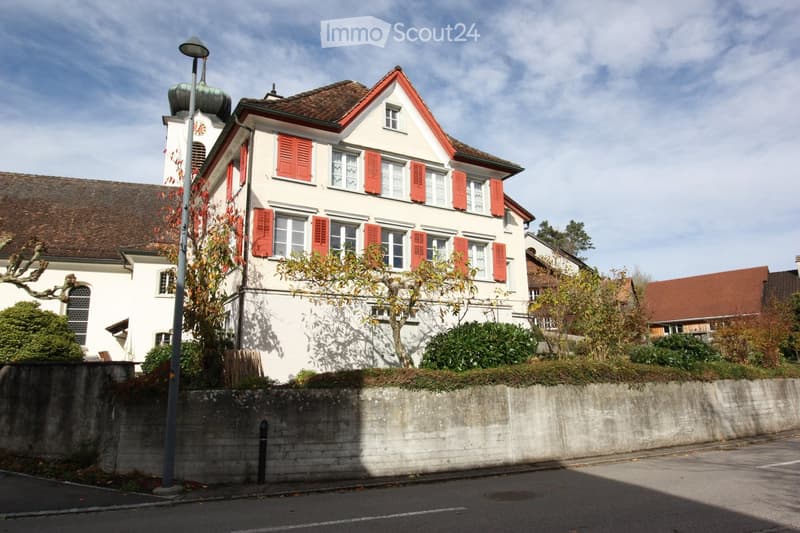 Two story house, white walls, red shutters, brown roof, street light, stone wall fence, small garden