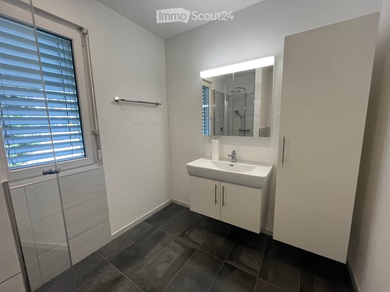 Modern bathroom with a white sink, large mirror with built-in lighting, dark tiled floor, and a window with blinds.