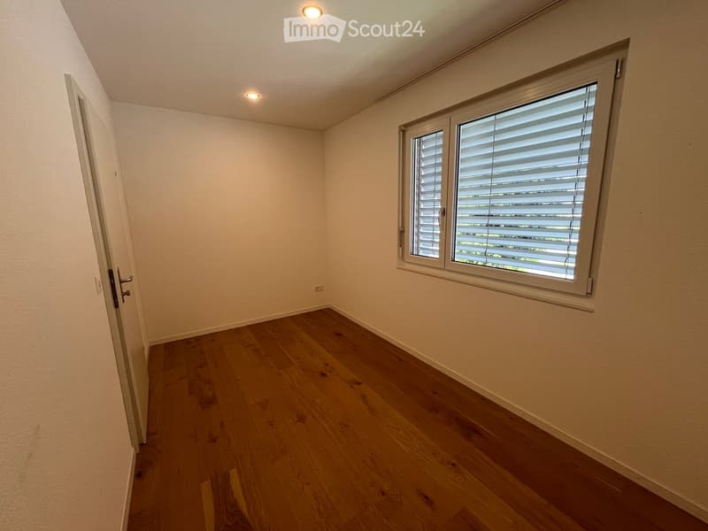 Empty room with wooden flooring, white walls, and a window with blinds. The room has recessed lighting and a door leading to another space.