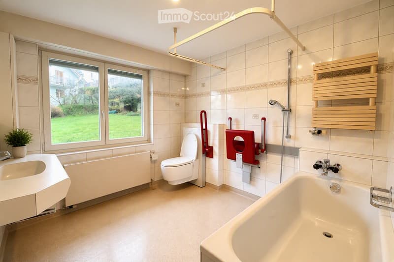 Modern bathroom with white tiles, bathtub, toilet, sink, and a large window overlooking a green area.