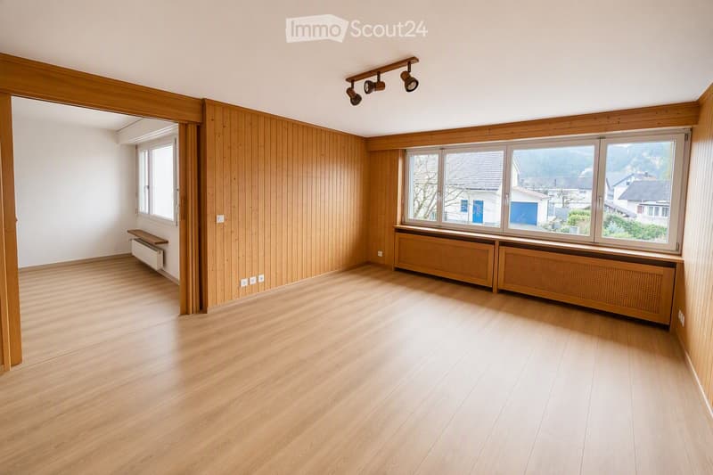 Empty room with wooden flooring, wooden panel walls, large windows with radiators, and track lighting.