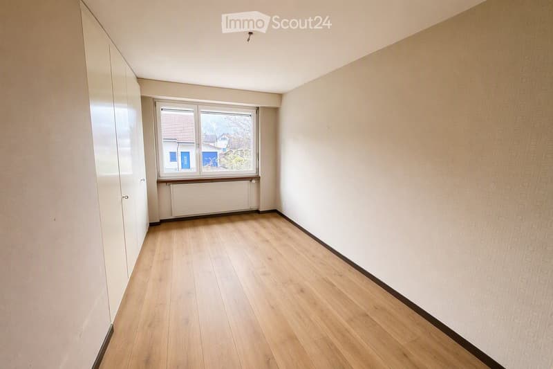 Empty room with light wooden flooring, white walls, and a large window offering a view of the outside. The room features a radiator below the window and a closet on the left side.