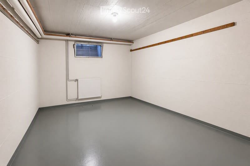 Empty room with white walls, grey floor, radiator, small window, and exposed pipes.