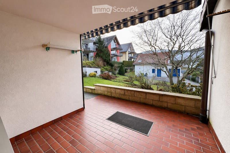 Balcony with red brick tiles, low stone wall, striped awning, and view of a landscaped garden and neighboring houses.