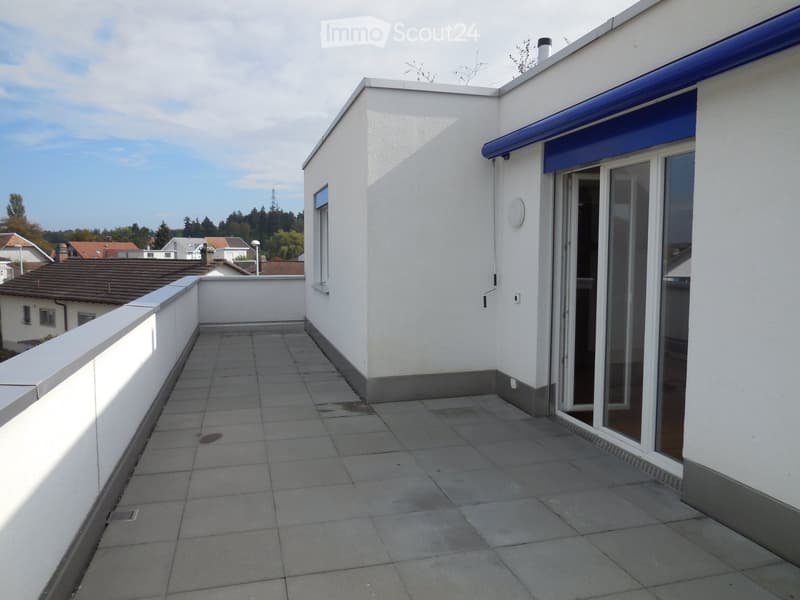 Roof terrace with tiled floor, white walls, and a sliding glass door leading inside. The area is clean and well-maintained, with a view of neighboring houses and trees.