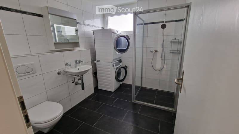 Modern bathroom with white tiles, black floor, toilet, sink, mirror, shower, and stacked washing machine.