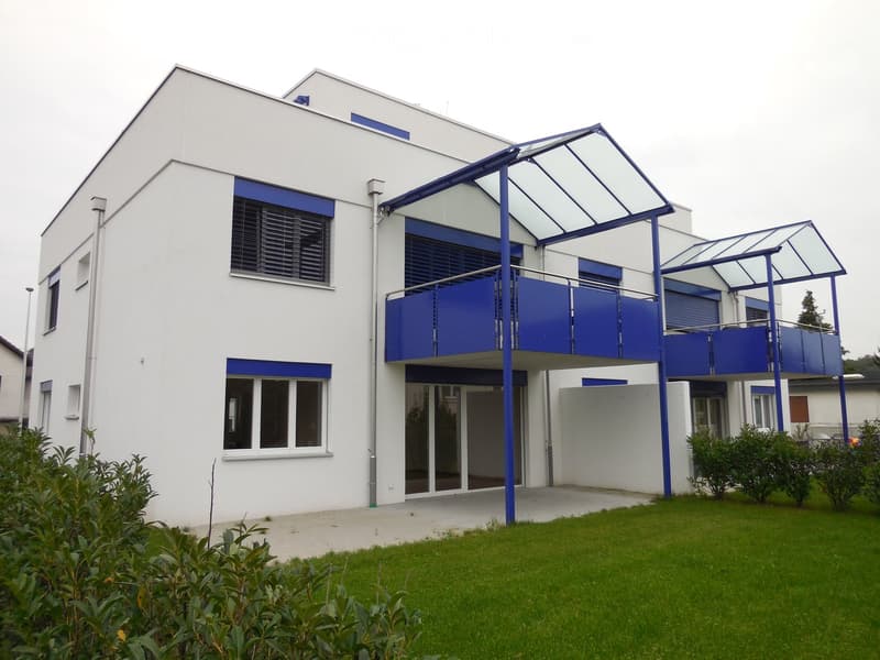 Modern two-story building with white walls, blue accents, large windows, and a covered entrance area with a glass roof.