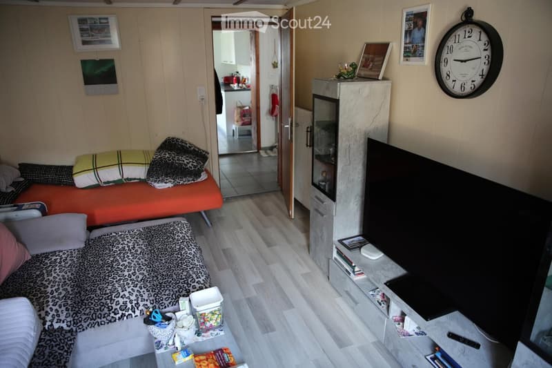 Cozy living space with a sofa, bed, TV, and small table. The room features wooden flooring and a view into an adjacent kitchen area.
