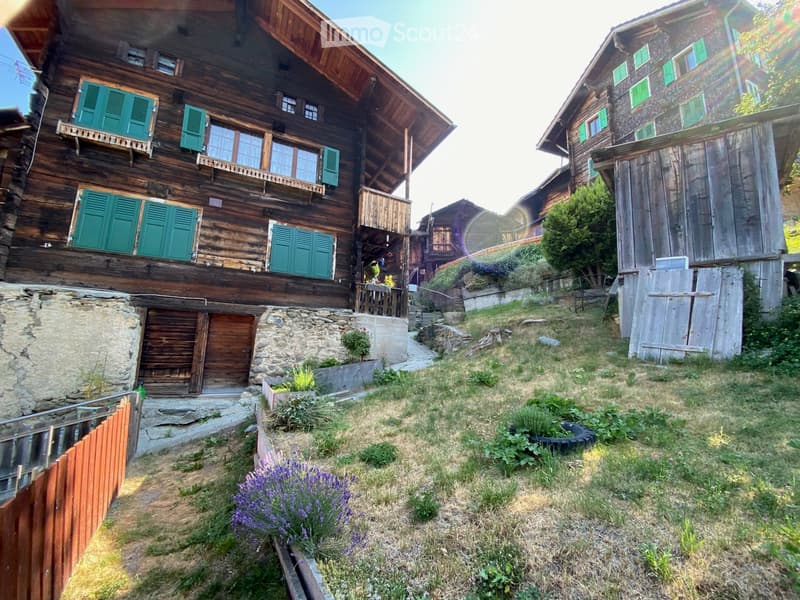 Traditional wooden chalet with green shutters, stone base, and wooden garage, surrounded by a grassy yard with lavender plants and a wooden fence.