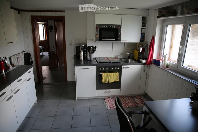 Modern kitchen with white cabinets, black countertops, and stainless steel appliances including an oven, microwave, and fridge. A small dining area with a table and chair is visible, along with a colorful rug on the floor.