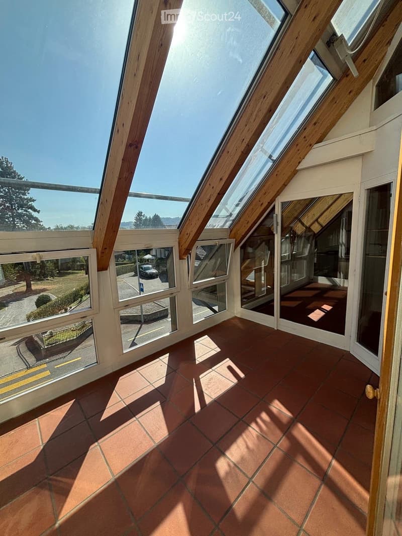 Bright, open attic space with wooden beams, large skylights, and terracotta tiled floor. The room has large windows offering views of the surrounding area, including a parking lot and greenery.