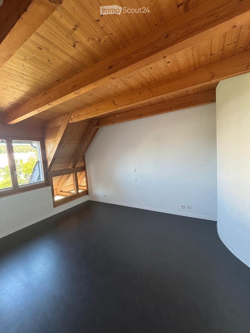 An empty attic space with exposed wooden beams, a dark grey floor, and a single window letting in natural light.
