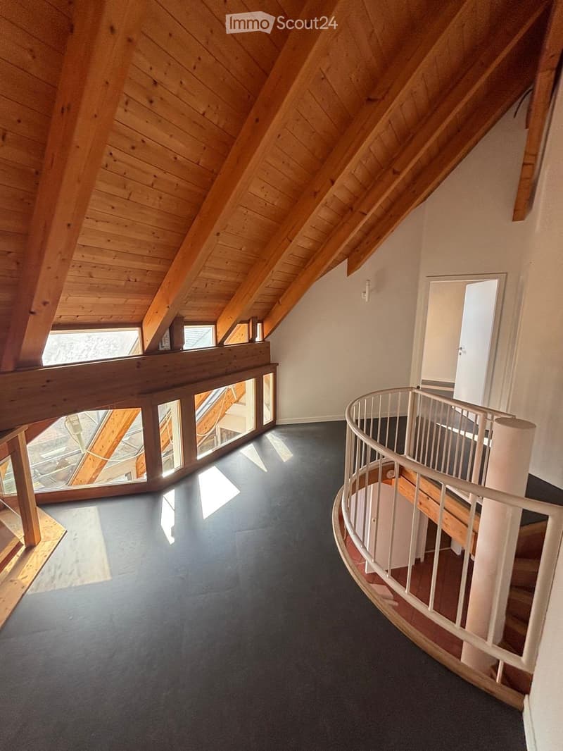 Spacious hallway with wooden ceiling beams, dark flooring, and a curved white staircase railing leading upwards.