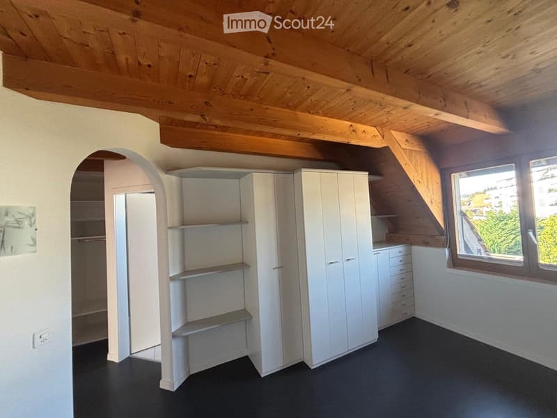 An unfurnished attic room with wooden beams, white built-in wardrobes, shelves, and large windows letting in natural light.