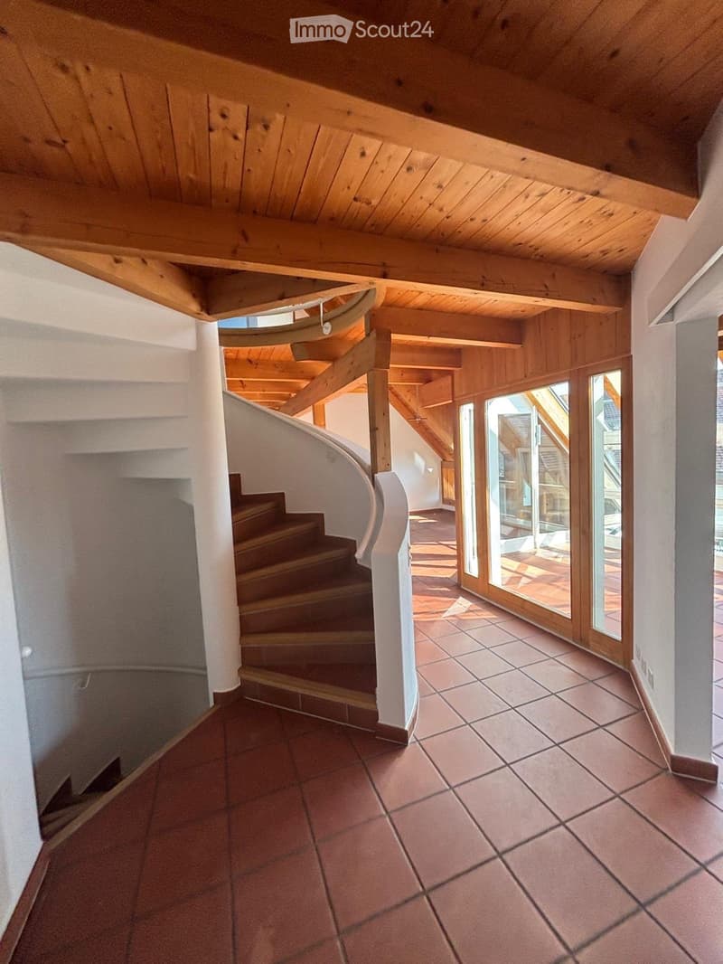Spacious hallway with exposed wooden beams, tiled floor, and a curved staircase leading upwards.