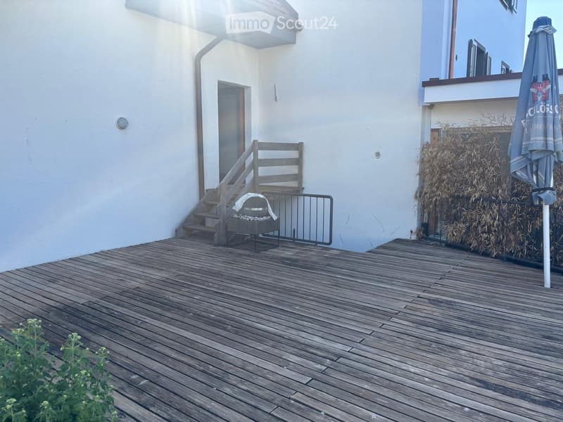 Wooden deck with stairs leading up, small seating area with a table and chairs, umbrella, and some plants.