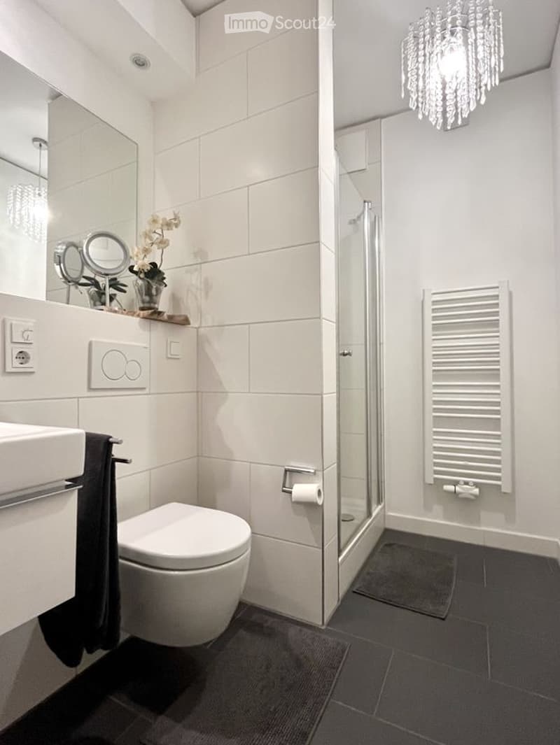Modern bathroom with white tiles, a toilet, sink, mirror, shower, radiator, and a decorative flower vase on the sink counter.