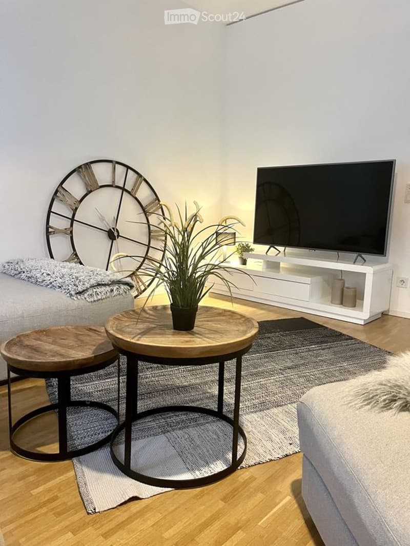 Modern living room with light-colored walls, wooden flooring, and a large window. Features a grey sofa, two wooden coffee tables, a television on a white stand, a large wall clock, and a potted plant on the coffee table.