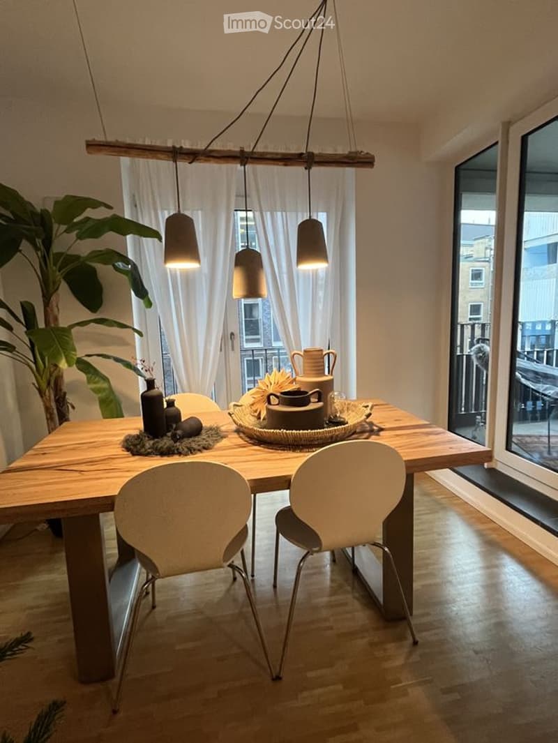 Modern dining area with wooden table, white chairs, hanging pendant lights, decorative items, and large windows offering a view.