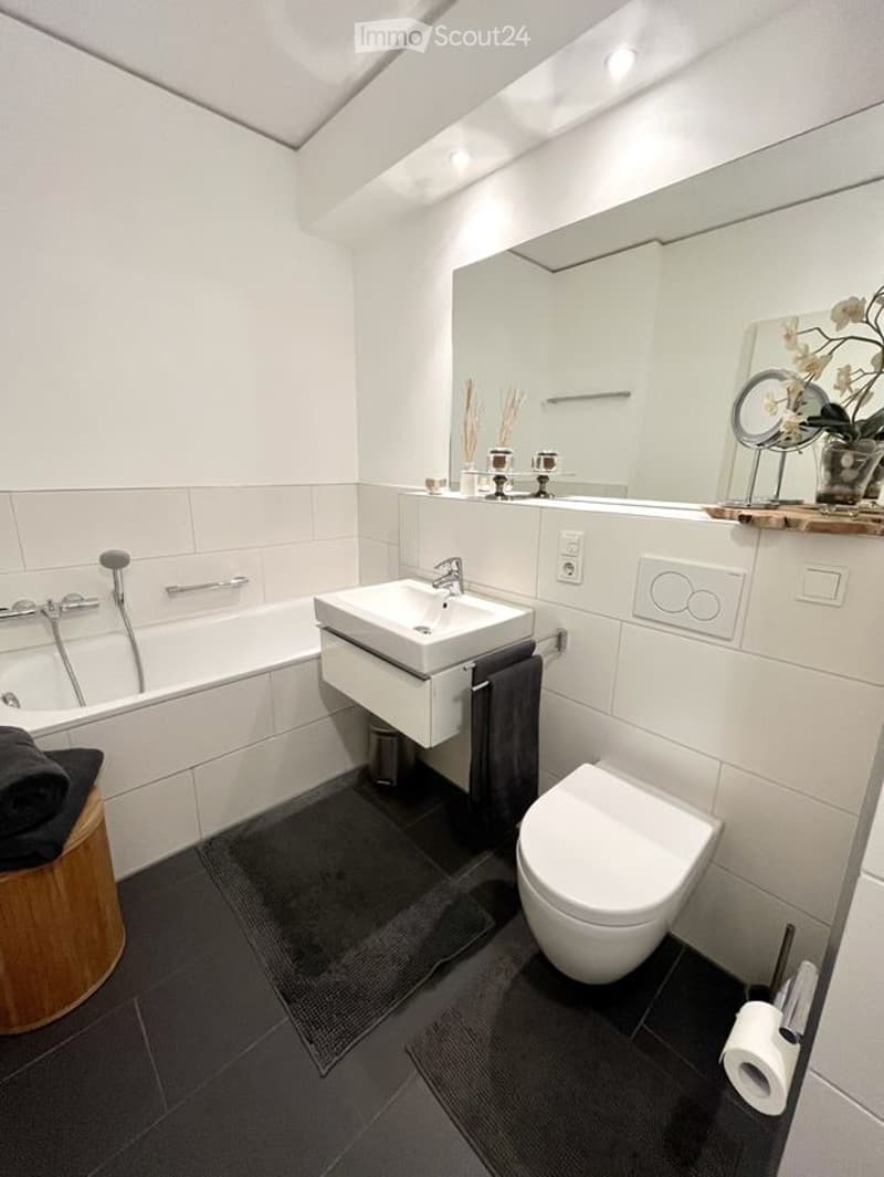 Modern bathroom with white tiles, a bathtub, a sink with a mirror, a toilet, and a wooden stool with a towel. The room features sleek fixtures and a dark grey floor.