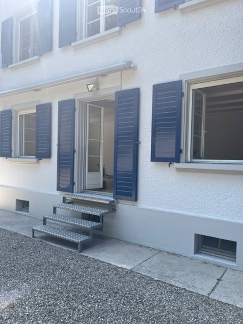 Exterior view of a two-story building with white walls, blue shutters, and a small staircase leading to the entrance door.