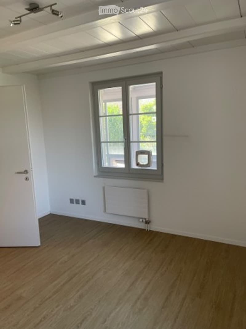 Empty room with wooden flooring, white walls, a single window with grey frame, radiator, and white door.