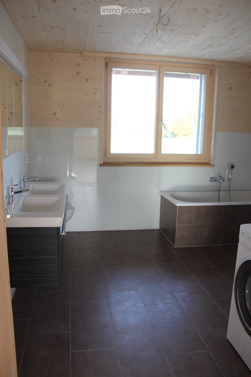 Modern bathroom with dark tiled floor, double sink, bathtub, and washing machine. Features wooden ceiling and large window with blinds.