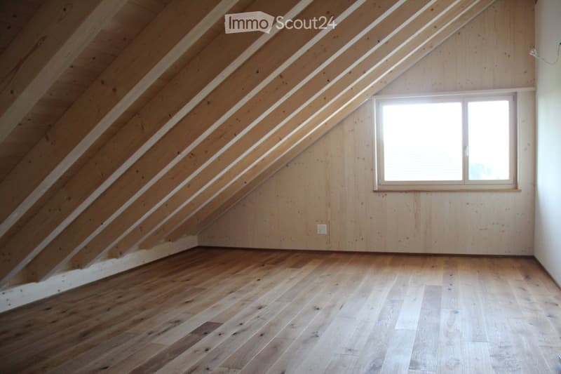 Empty attic space with wooden walls, slanted wooden ceiling, and a single window letting in natural light. The floor is made of light-colored wooden planks.