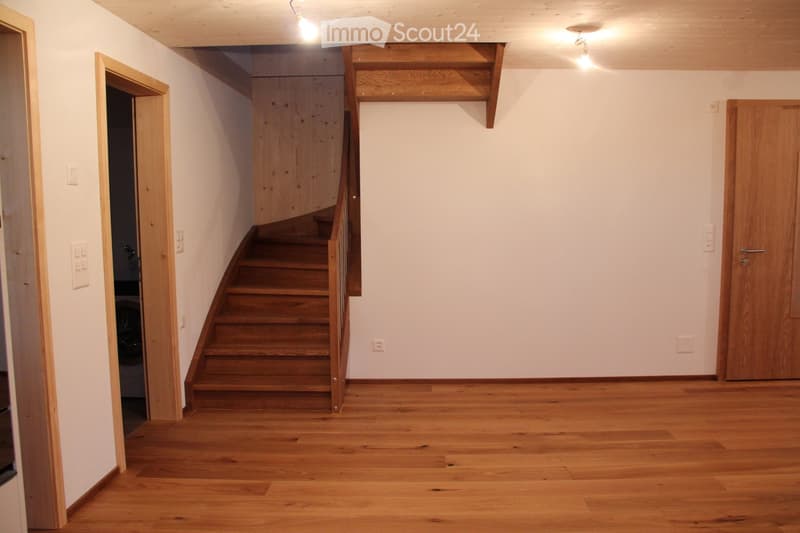 Empty hallway with wooden flooring, white walls, and a staircase leading upwards. There are doors on both sides and light fixtures on the ceiling.