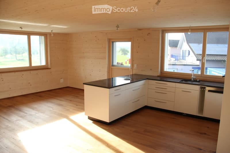 Modern kitchen with light wood walls, white cabinets, black countertops, and a central island. Large windows provide natural light.