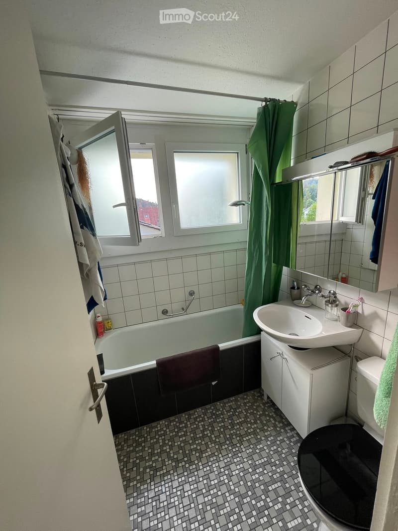Bathroom with a bathtub, sink, toilet, mirror, and tiled walls and floor. A green shower curtain is visible, and a towel hangs on the side of the bathtub.