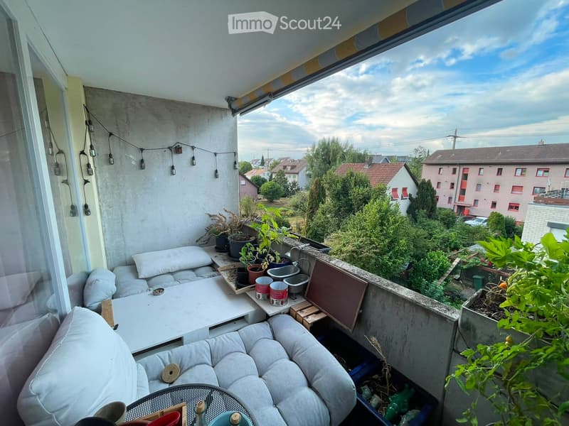Balcony with a white sofa, small table, and several potted plants, overlooking a residential area with trees and houses.
