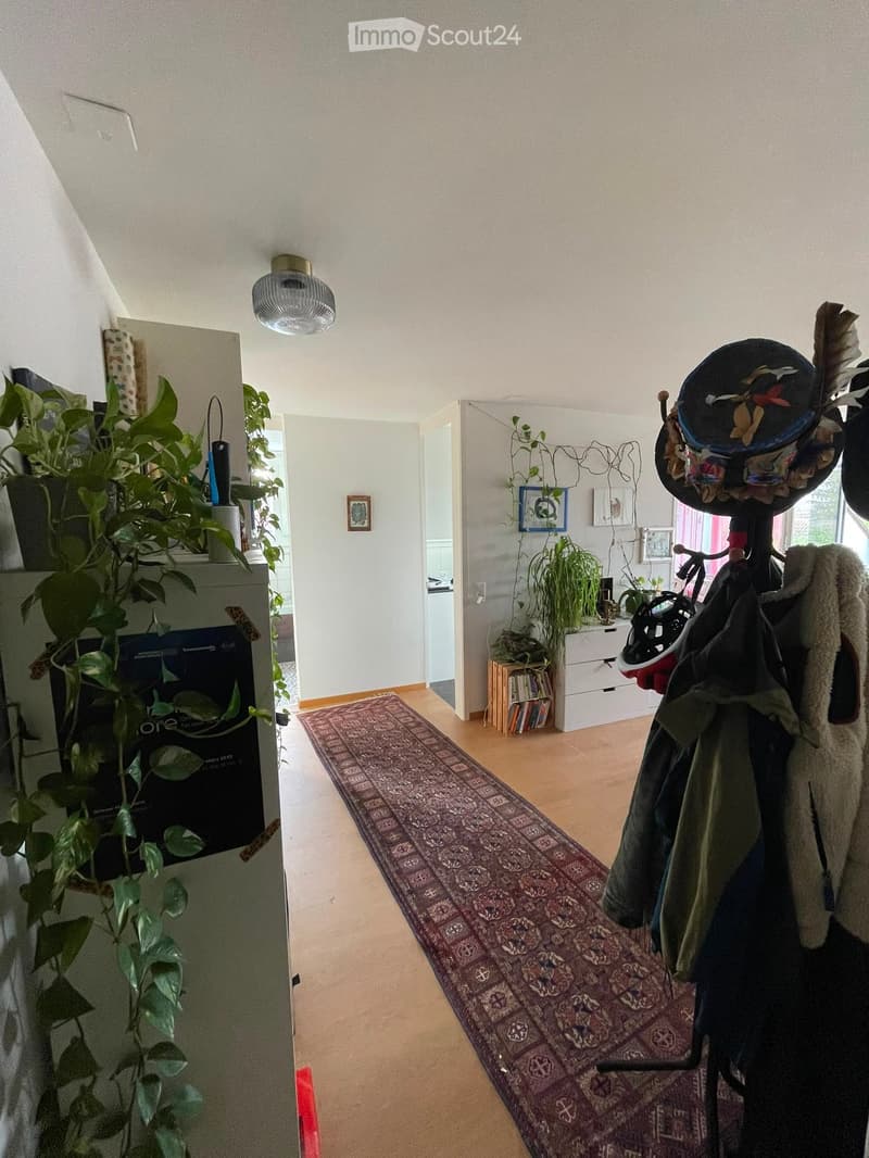 Bright hallway with wooden flooring, a patterned rug, coat rack with jackets, and potted plants on both sides.