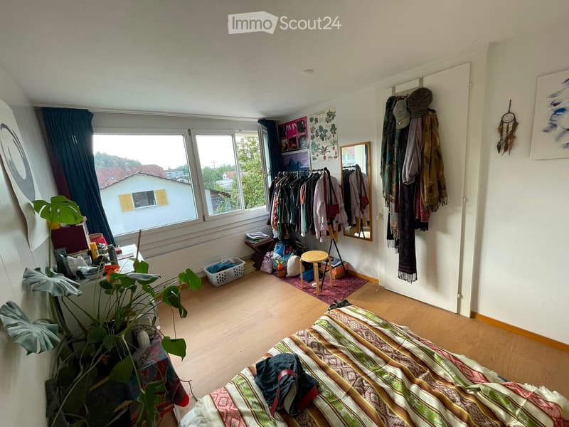 Cozy bedroom with wooden flooring, large windows offering a view, a bed with a colorful striped blanket, a small wooden stool, a clothing rack, a mirror, and a dreamcatcher on the wall.