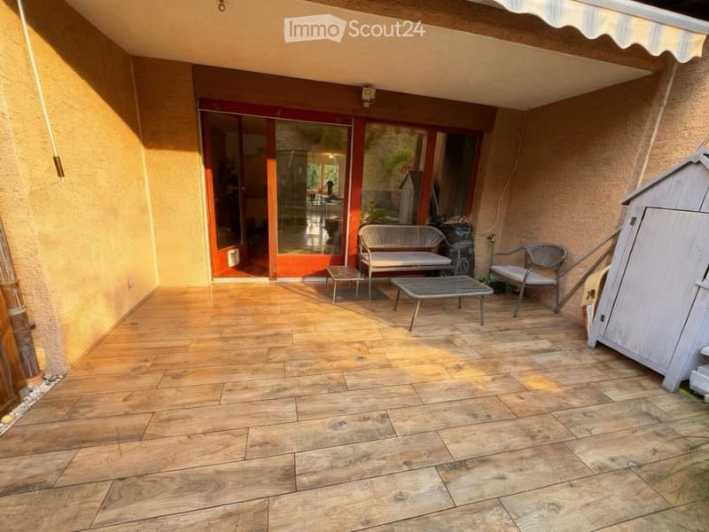 Balcony with wooden flooring, a bench, a small table, and a chair, connected to a room with large glass doors.