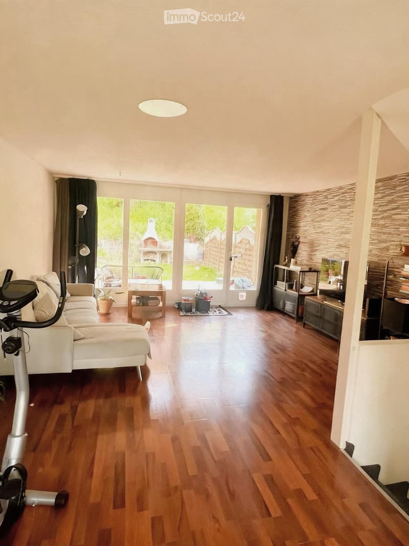 Bright living room with wooden flooring, large windows, a white sofa, exercise bike, small table with chairs, and a shelf with decorative items.
