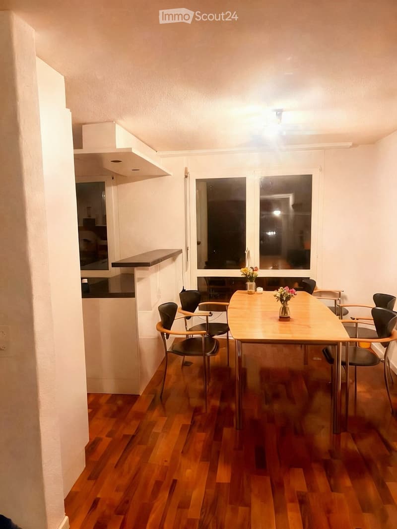Dining area with wooden table, chairs, and vases on the table. The room has wooden flooring and white walls.