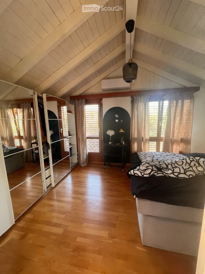 Bright attic bedroom with wooden flooring, slanted wooden ceiling, large windows with curtains, a bed with black and white bedding, a wardrobe, and a small table with a lamp.